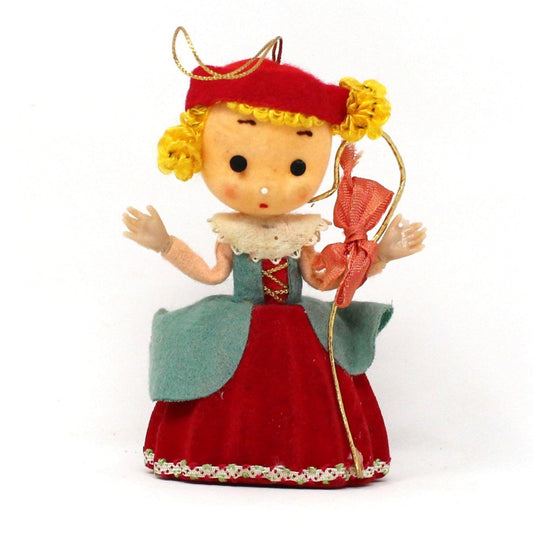 A vintage flocked Little Bo Peep Christmas ornament with yarn hair and plastic hands, wearing a red dress and hat, with a gold staff with red bow.
