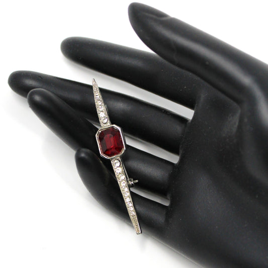 A vintage bar brooch featuring a geometric design with a large ruby red stone and pave rhinestones in silver-tone metal displayed on a black hand mannequin.