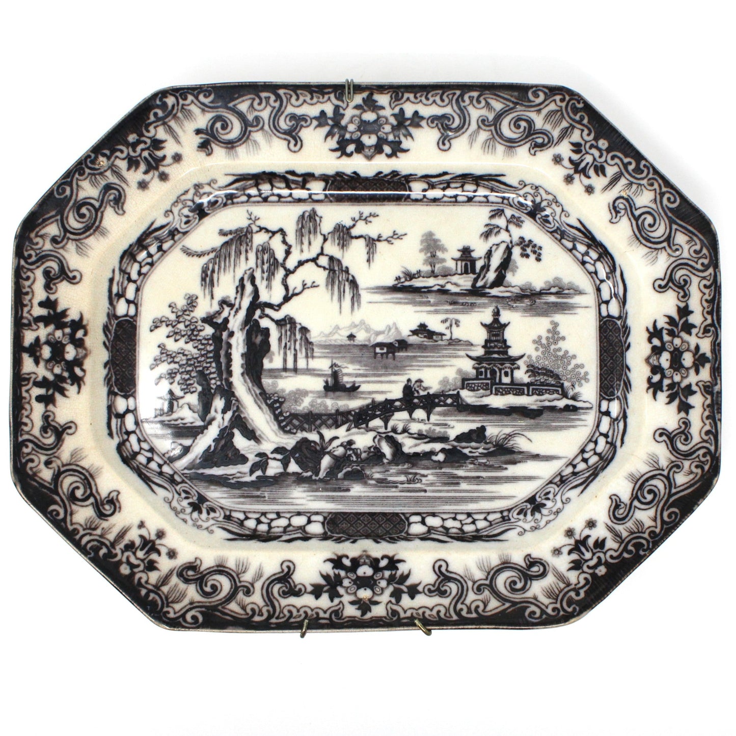 Antique serving platter with a black transferware design featuring a Chinese pagoda, willow trees, a wooden bridge, and mountains. The platter has an intricate border design with swirls and flowers.