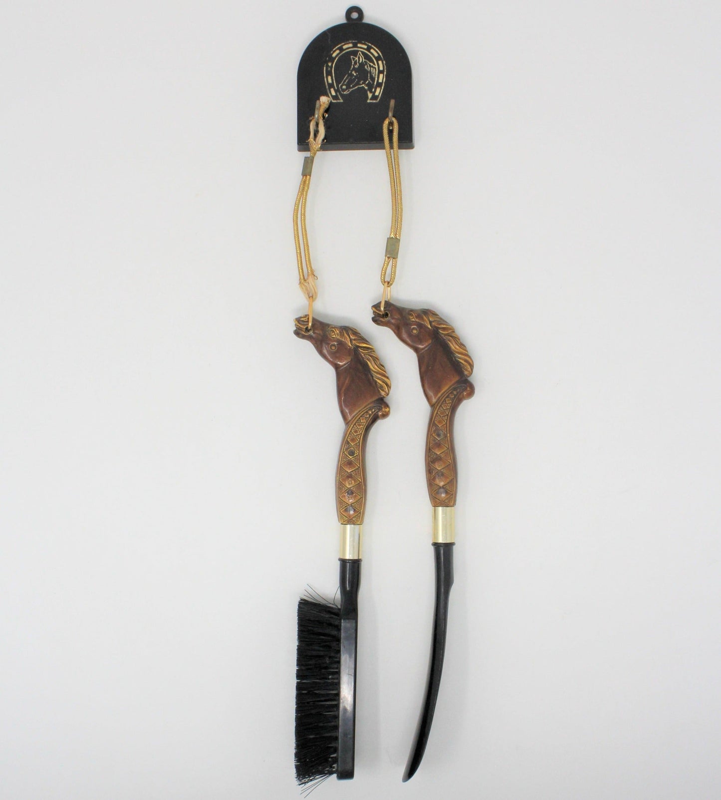 A vintage equestrian shoe horn and brush set with horse head handles, with black plastic hanger and two hooks.