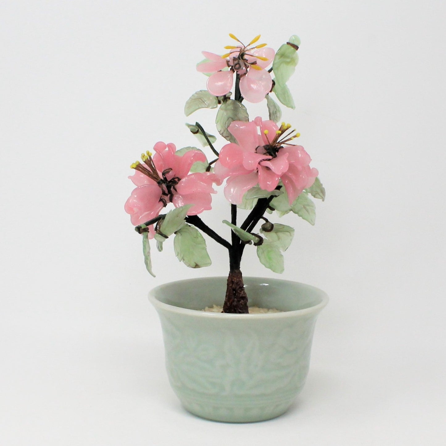 A jade glass bonsai tree sculpture in a celadon planter with pink flowers and green leaves.