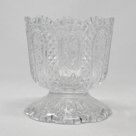 A vintage Fostoria clear glass candle holder with a Daisy and Diamond pattern, featuring a footed base and a pedestal base, with the brand name 'AVON' embossed on the bottom.