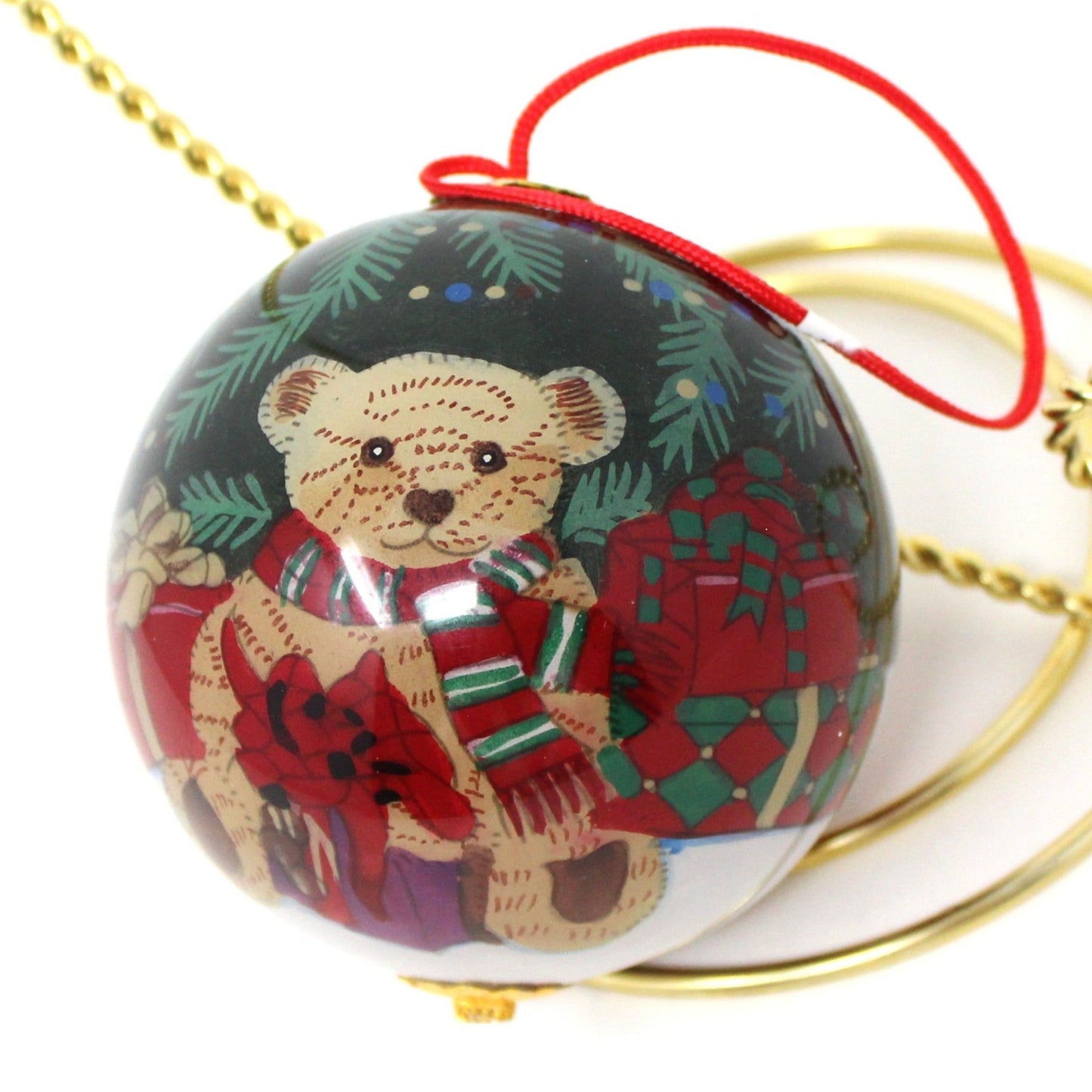 A reverse painted teddy bear Ganz Christmas glass ball ornament. Bear wearing a scarf and gifts on a red background, with a gold metal cap and red satin cord for hanging.