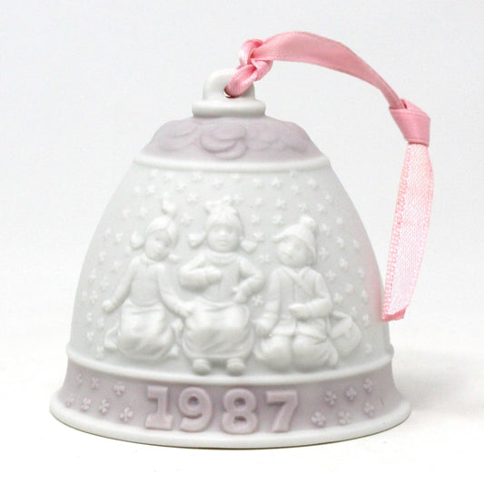 A pink and white porcelain Lladro Christmas bell ornament with children playing in the snow, and a gold metal chain on top. The year 1987 is embossed on the bell.