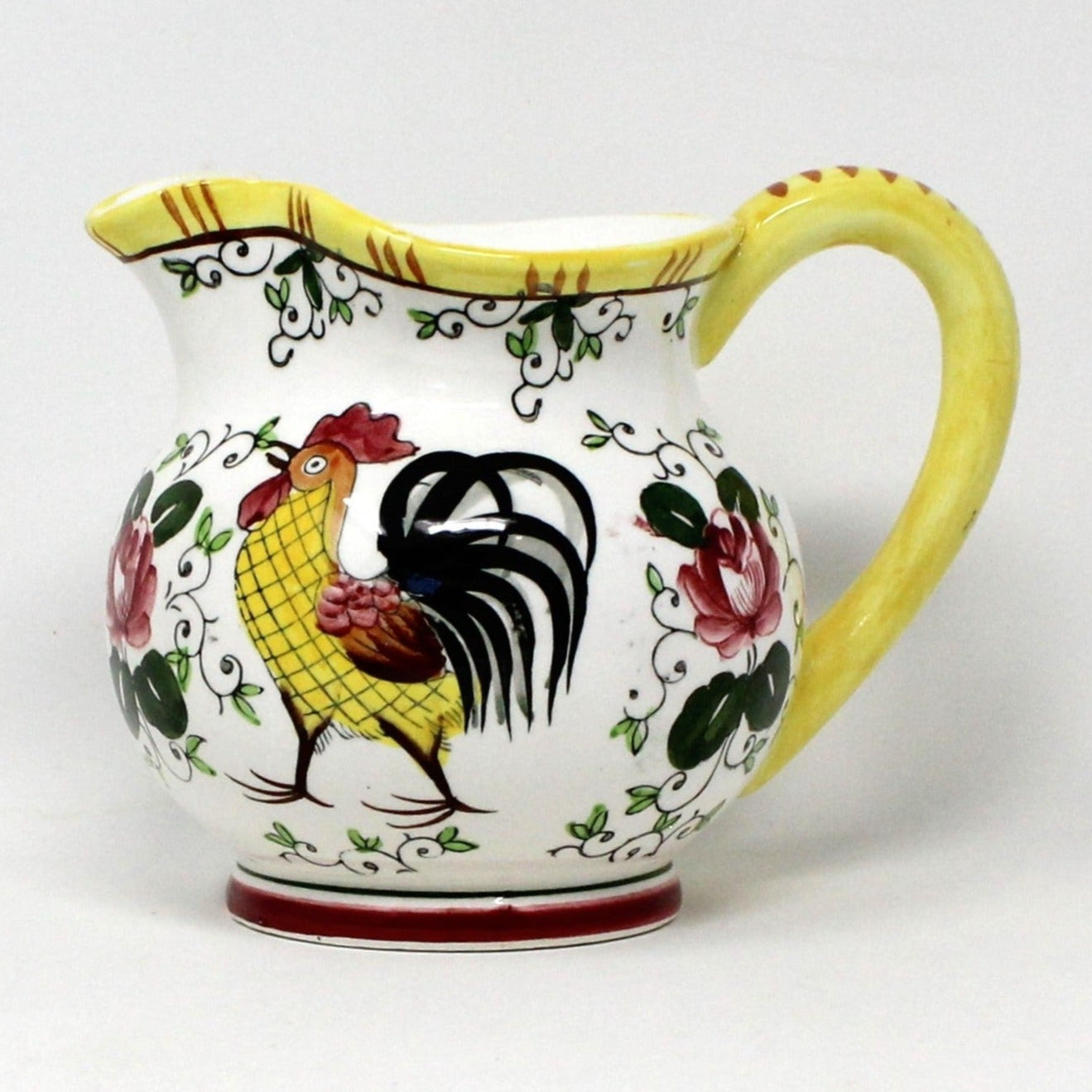 A ceramic creamer / pitcher with a yellow handle and a vibrant rooster and floral pattern featuring large red roses, bordered in yellow.
