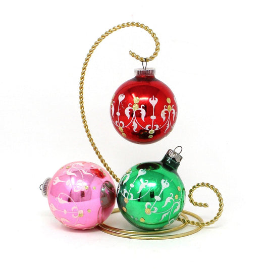 Set of three vintage Christmas ornaments made of glass with red, green, and pink colors, featuring white stencil designs and gold glitter, and plastic caps marked 'Made in Austria'.