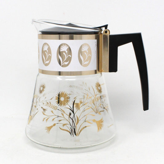 A vintage clear glass coffee carafe with a gold flowers and wheat pattern, featuring a black lid and handle, and metal accents.