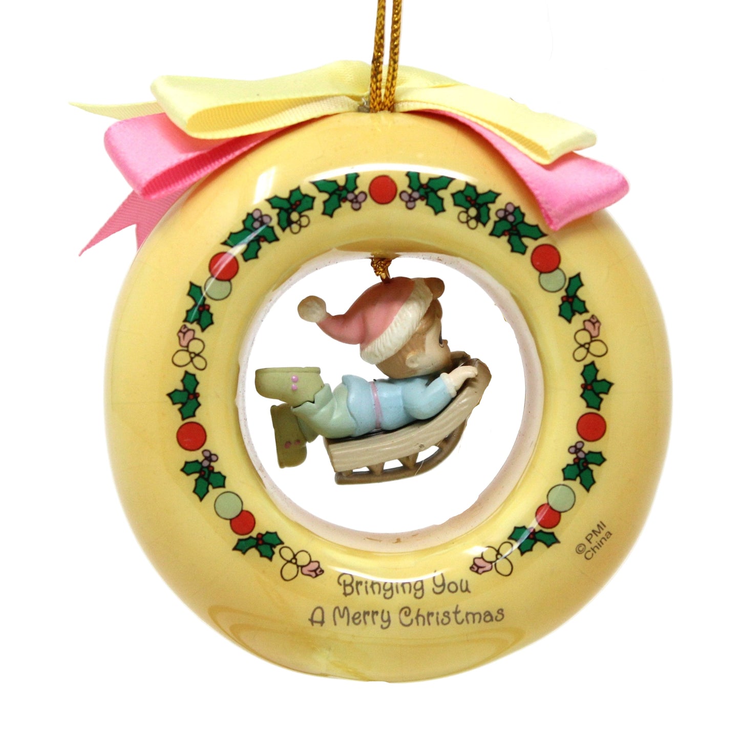 A Precious Moments vintage Christmas ornament featuring a boy on a sled dangle charm, with a yellow base, holly decorations, and a gold cord.