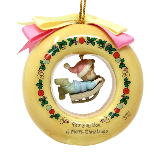 A Precious Moments vintage Christmas ornament featuring a boy on a sled dangle charm, with a yellow base, holly decorations, and a gold cord.