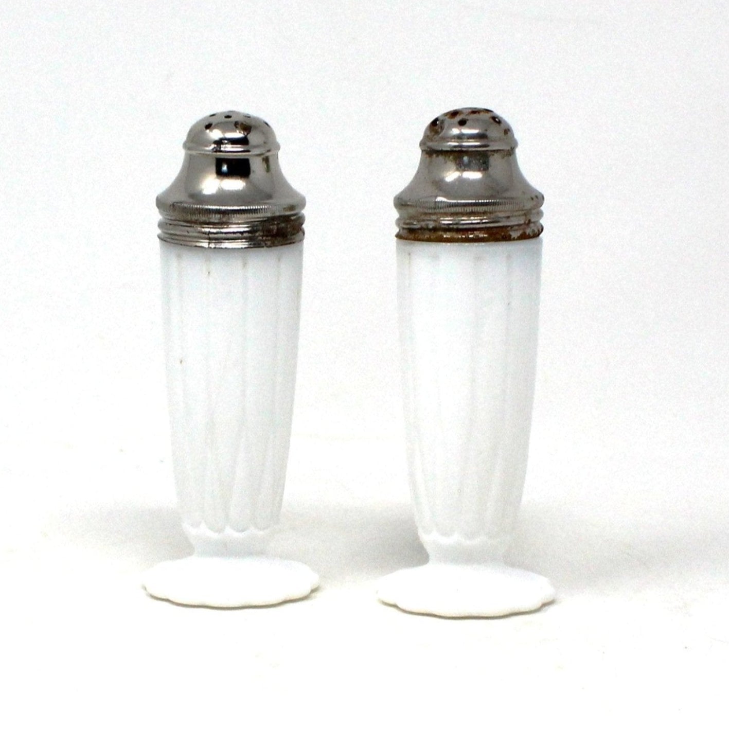 A pair of vintage Hazel Atlas milk glass salt and pepper shakers with metal lids, featuring a white exterior and a hairpin design on the footed base.