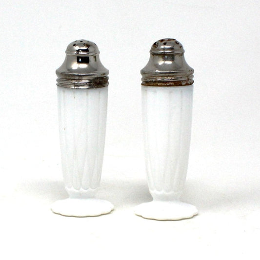 A pair of vintage Hazel Atlas milk glass salt and pepper shakers with metal lids, featuring a white exterior and a hairpin design on the footed base.