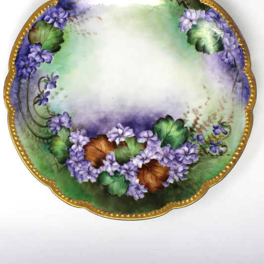 close up view of an antique scalloped Limoges France porcelain plate with a hand-painted floral design featuring purple flowers, green & brown leaves, and a gold moriage rim.
