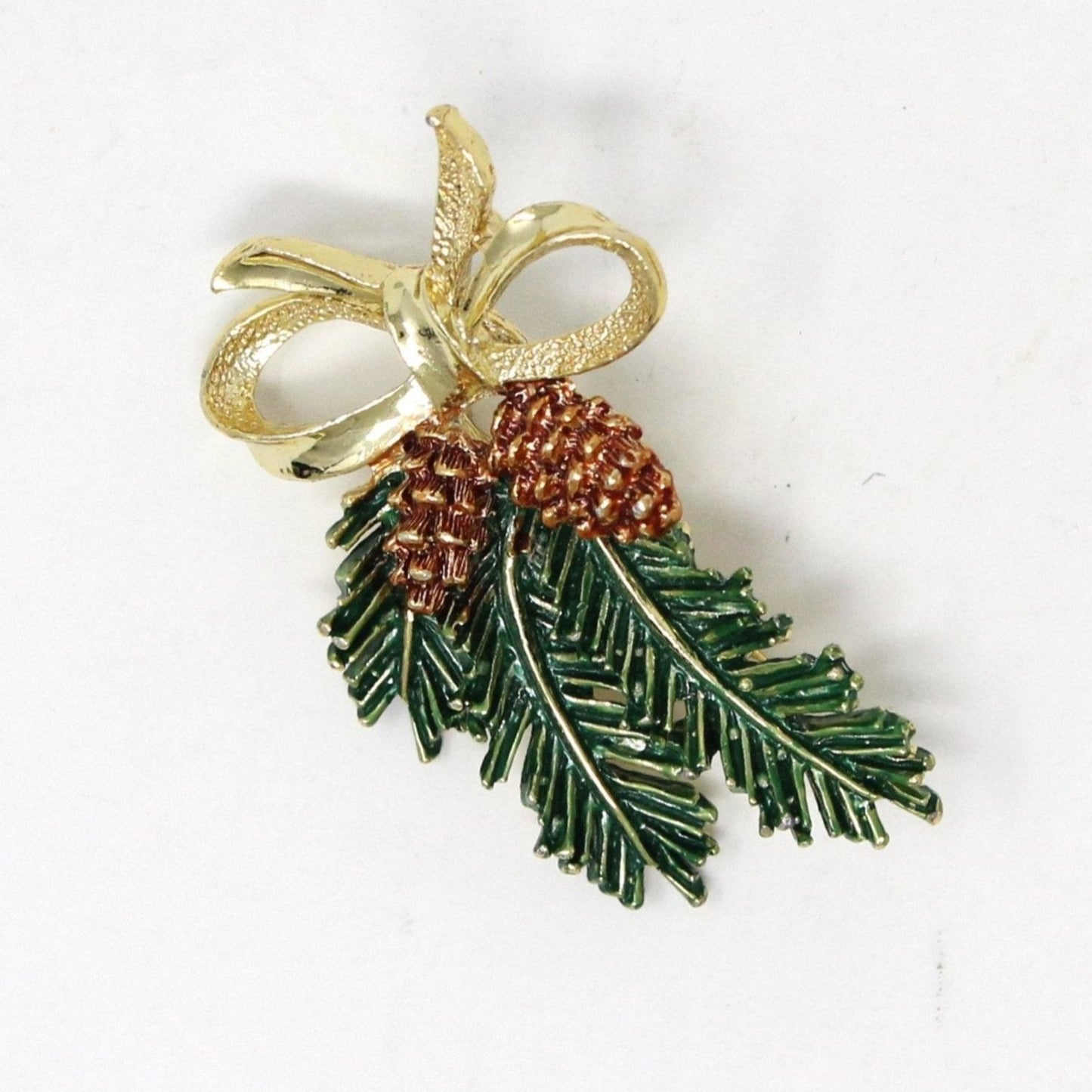 A vintage gold-tone metal brooch with enamel detailing, featuring two pine cones and green pine branches tied with a gold bow.