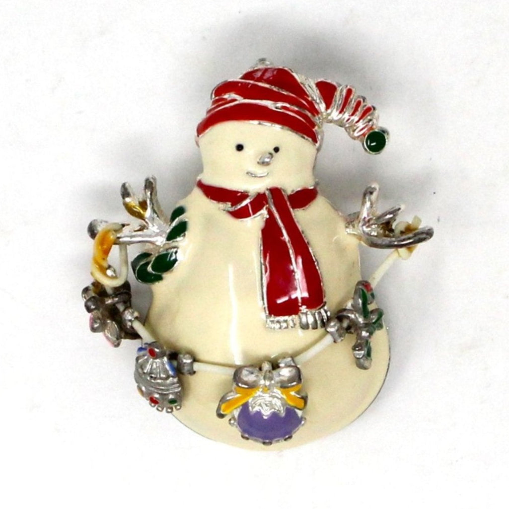A silver-tone metal brooch featuring a white enamel snowman with a red hat and scarf, holding dangle ornaments, and a Christmas tree, candy cane, ornament, and bell in bright enamel colors.