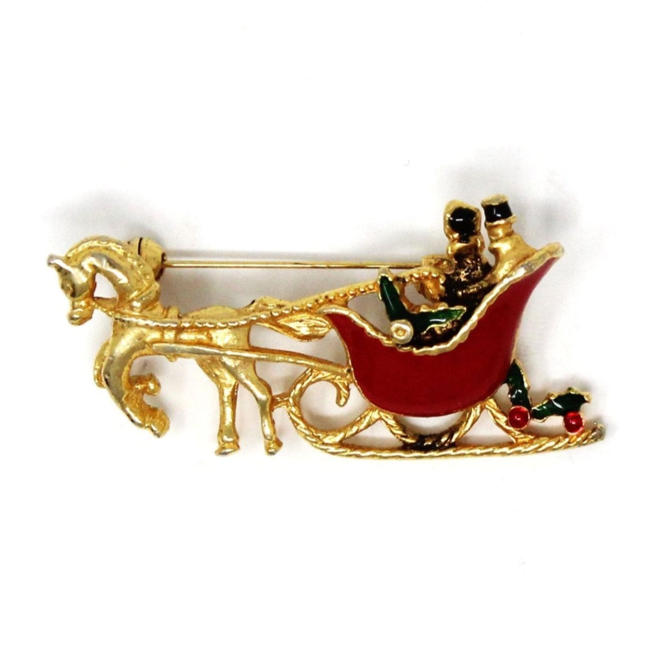 A vintage gold-tone metal brooch featuring a horse-drawn sleigh with red enamel details, including passengers and decorations in the form of green holly and red berries.