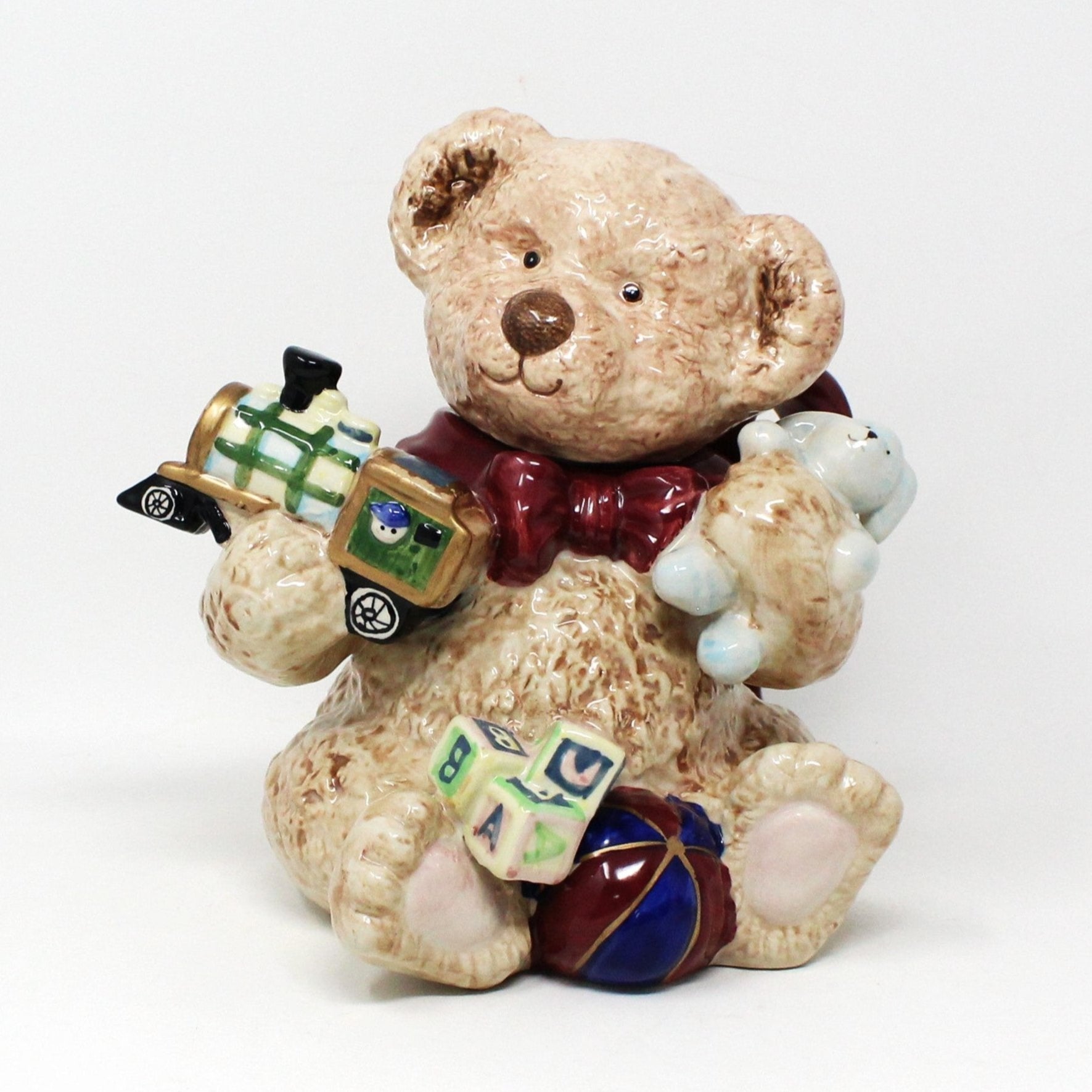 A ceramic teapot shaped like a teddy bear holding various Christmas toys such as a train, blocks, a ball, and a bunny.