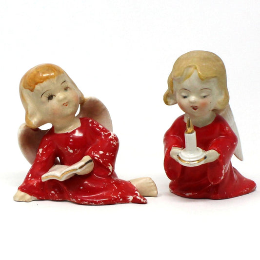 A pair of hand-painted porcelain figurines depicting Christmas angels, one angel reading a book and the other holding a candle, both in red and white colors.