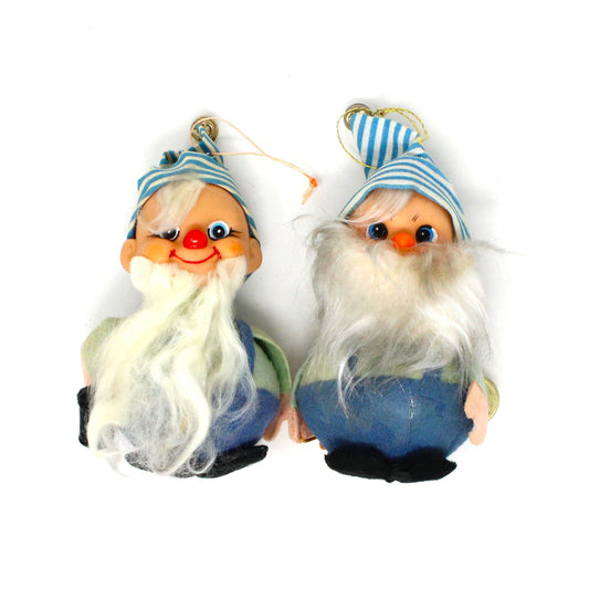 Two vintage Christmas ornaments featuring jolly elves with long beards, one wearing blue overalls and a striped hat with a bell, and the other with a similar outfit but without the mallet.