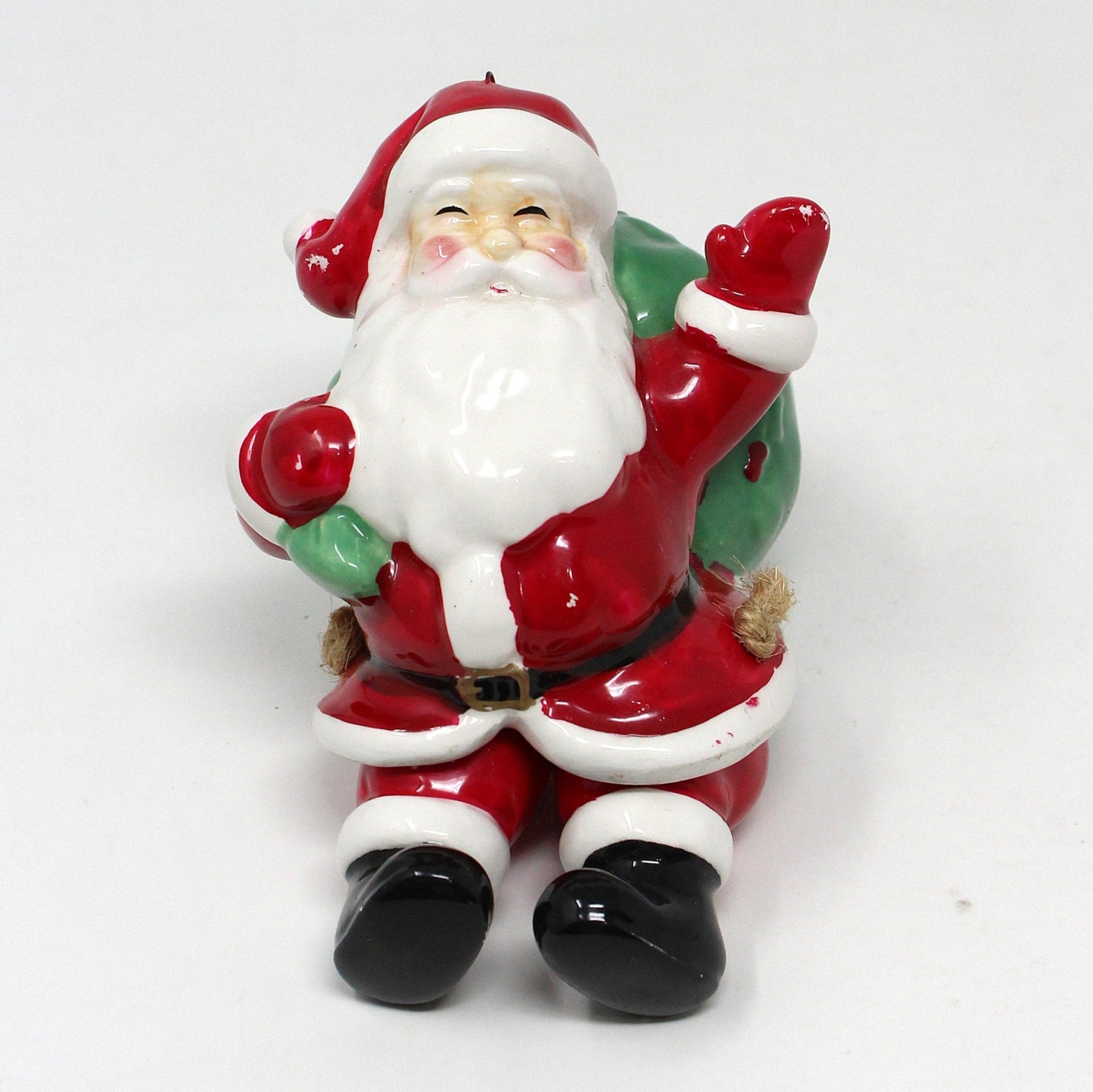 A ceramic ornament depicting Santa Claus with a large gift sack and dangling movable legs.