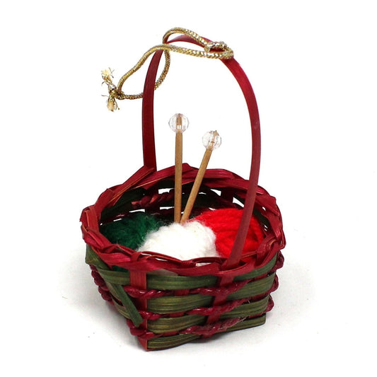 A vintage Christmas ornament featuring a red and green wicker basket filled with yarn and knitting needles, with a gold cord for hanging.