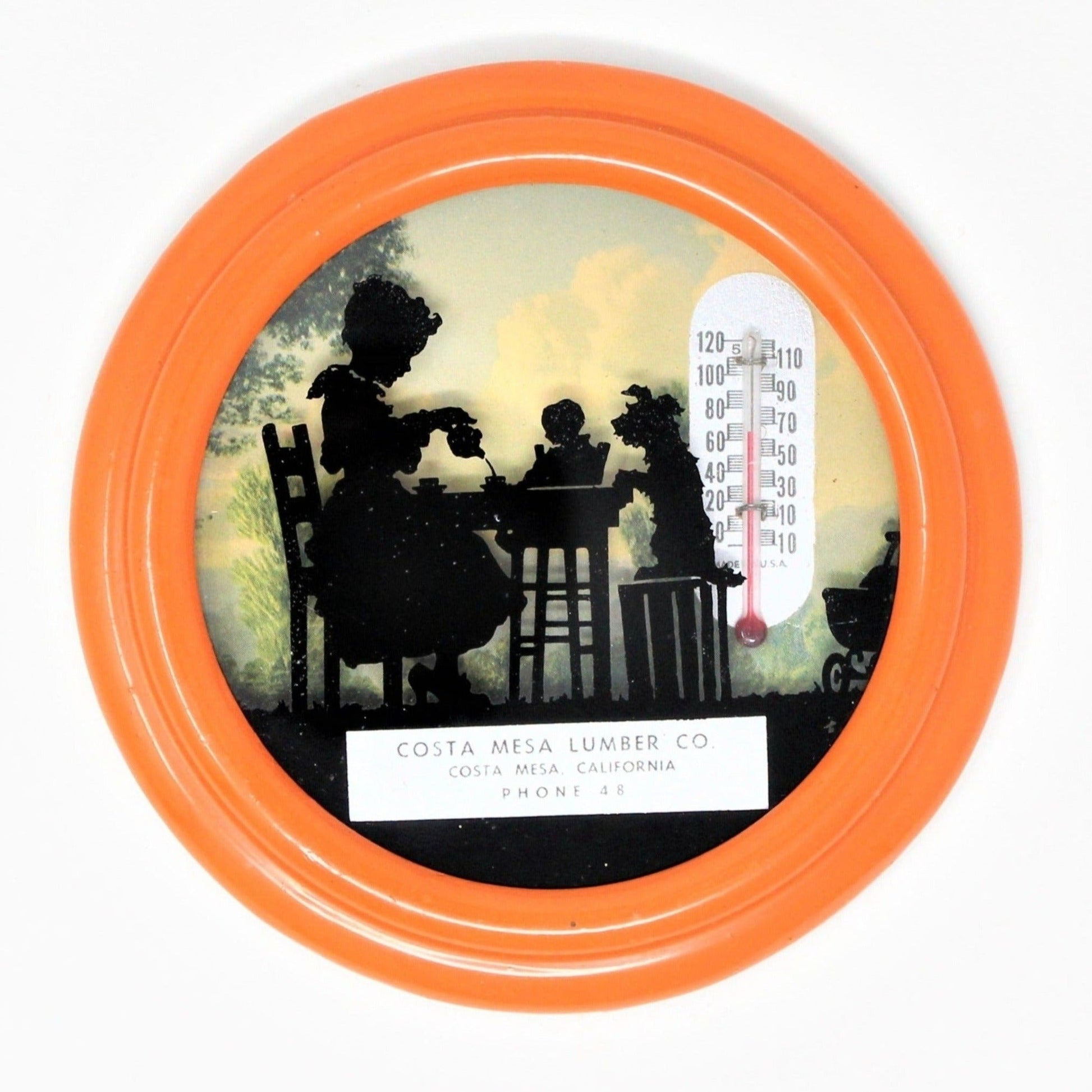 Antique advertising thermometer with a reverse painted silhouette showing a child playing 'tea time' with a doll and dog. The thermometer has a round, orange metal frame. Advertisement for Costa Mesa Lumber Co, California.
