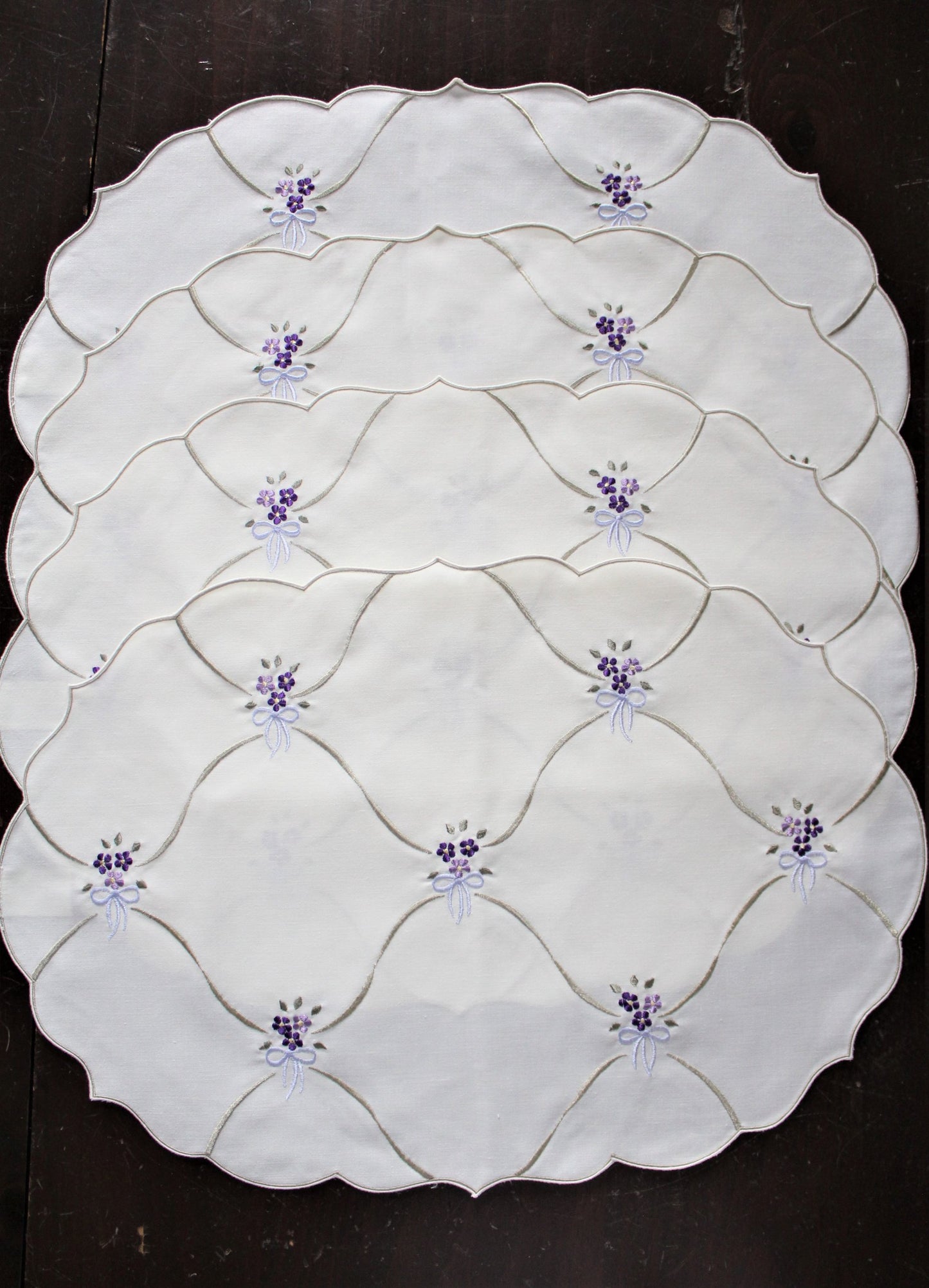 A set of four oval placemats with embroidered purple floral bouquets and blue bows on an eggshell white background.