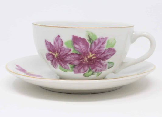 A vintage white porcelain teacup with a purple orchid pattern and gold detailing, accompanied by a matching saucer with a smaller orchid design, both stamped 'B-954 Japan'.