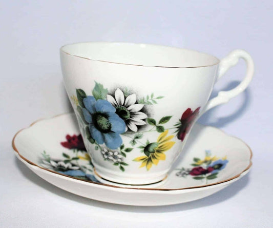 A bone china teacup and saucer with a vintage floral pattern in blue, red, yellow, and white, featuring gold ruffled details and a gold rim.