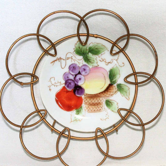 A decorative porcelain plate with a hand-painted fruit design, encased in a bronze-colored wire frame, featuring a basket of grapes, an apple, and a pear with leaves and tendrils.