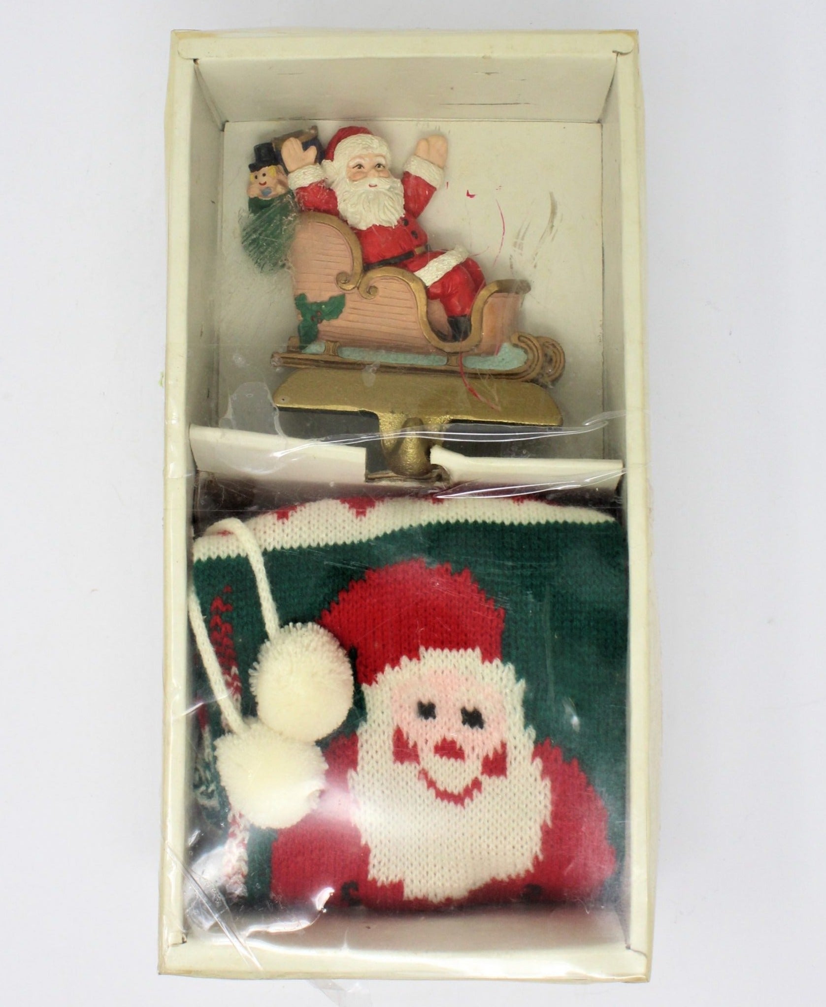 A vintage Santa Claus-themed stocking hanger with a sleigh and Christmas trees, accompanied by a knitted stocking featuring Santa and pom-poms.