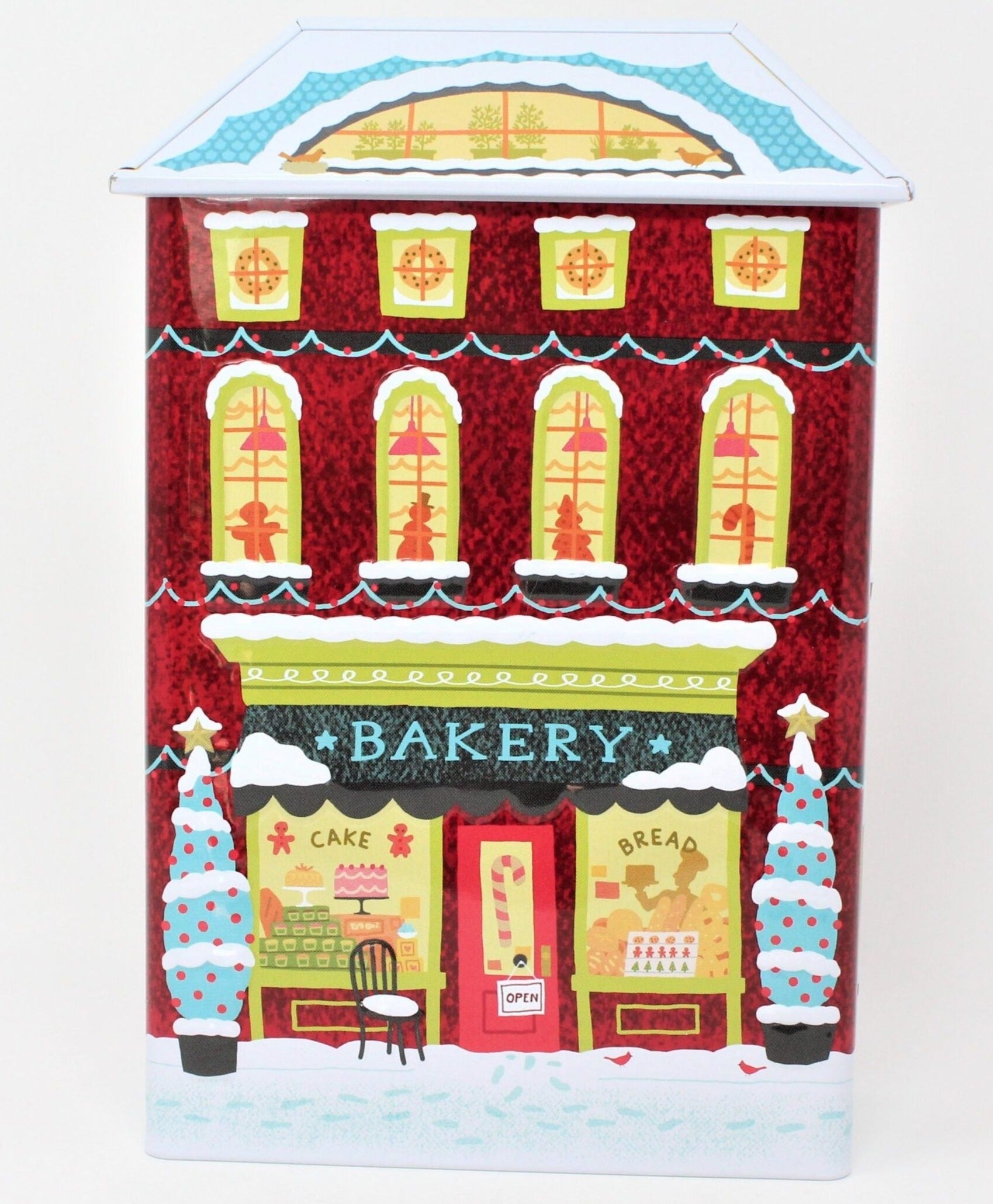 A collectible Christmas-themed cookie tin shaped like a red brick apartment building with a bakery shop on the bottom floor, decorated with festive designs and people engaging in holiday activities.