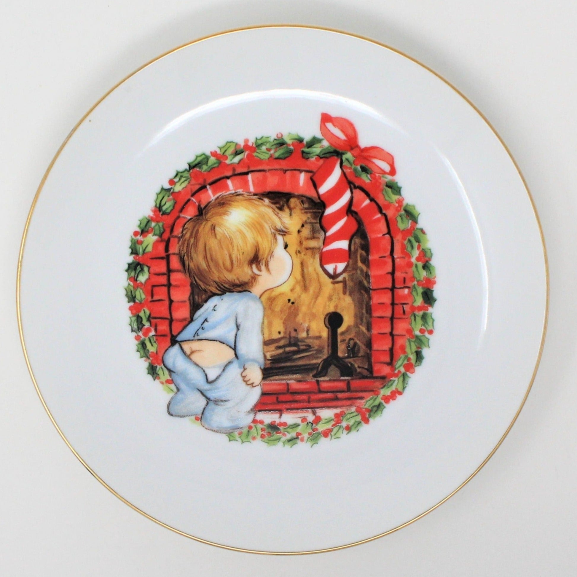 A decorative porcelain plate featuring a vintage Christmas design with a little boy by a fireplace, surrounded by holiday decorations and a red and white color scheme.