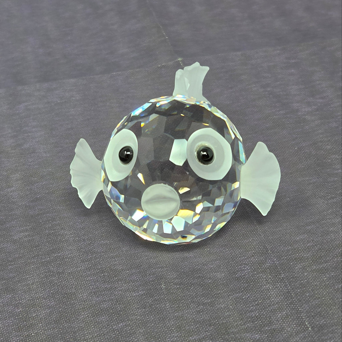 A clear faceted crystal fish ornament with white painted details and black bead eyes, placed on a textured gray fabric background.