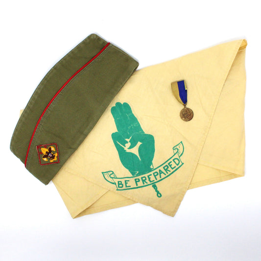 A collection of vintage Boy Scouts of America memorabilia including a yellow neckerchief with text 'Be Prepared' and a three-finger salute, a green mid-century hat with a badge, and a gold-toned Cub Scout Rocket Derby Medal with blue and yellow ribbon.