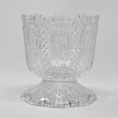 A vintage Fostoria clear glass candle holder with a Daisy and Diamond pattern, featuring a footed base and a pedestal base, with the brand name 'AVON' embossed on the bottom.