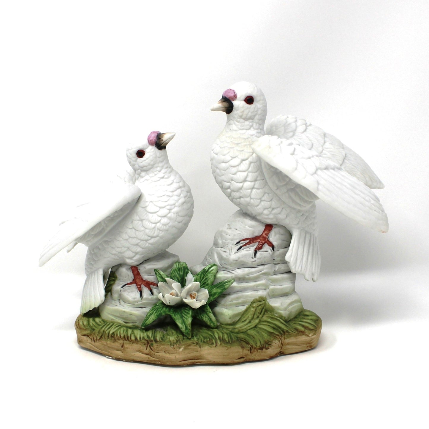 A decorative vintage figurine of two white doves standing on rocks, with green leaves and small white flowers.