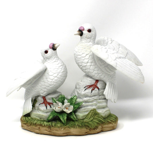 A decorative vintage figurine of two white doves standing on rocks, with green leaves and small white flowers.