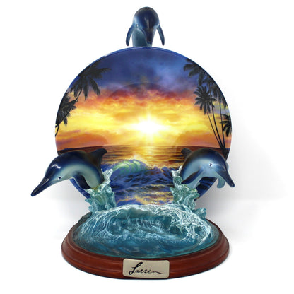 A decorative figurine of three dolphins jumping from blue ocean waves under a colorful sunset sky, mounted on a round wooden base with a small plaque.