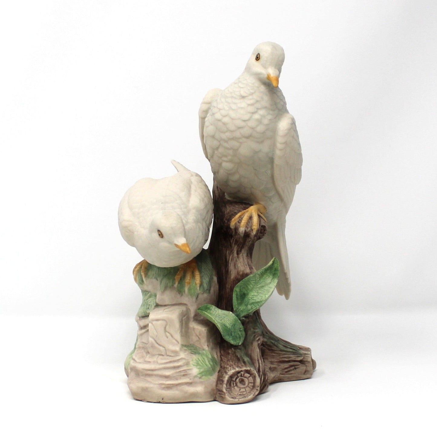 A ceramic figurine of two white doves perched on a brown tree trunk with green leaves, set against a plain white background.