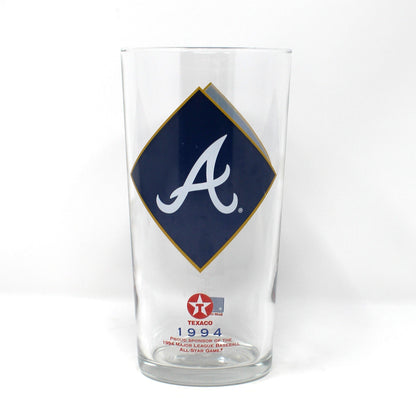 A clear glass beer pint glass featuring the Atlanta Braves logo, with a navy blue diamond on the front displaying a white script letter 'A' and a gold outline, plus a small red and white Texaco star logo near the base.