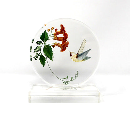 A round clear glass coaster featuring a hand-painted design of a hummingbird and green leaves with red-orange flowers, displayed on a clear acrylic stand against a white background.