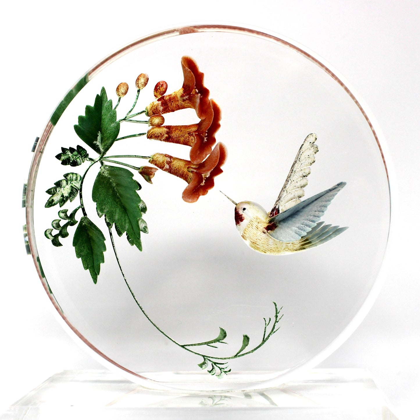 A round clear glass decorative plate featuring a painted illustration of a hummingbird and red-orange flowers with green leaves, displayed on a clear acrylic stand against a white background.