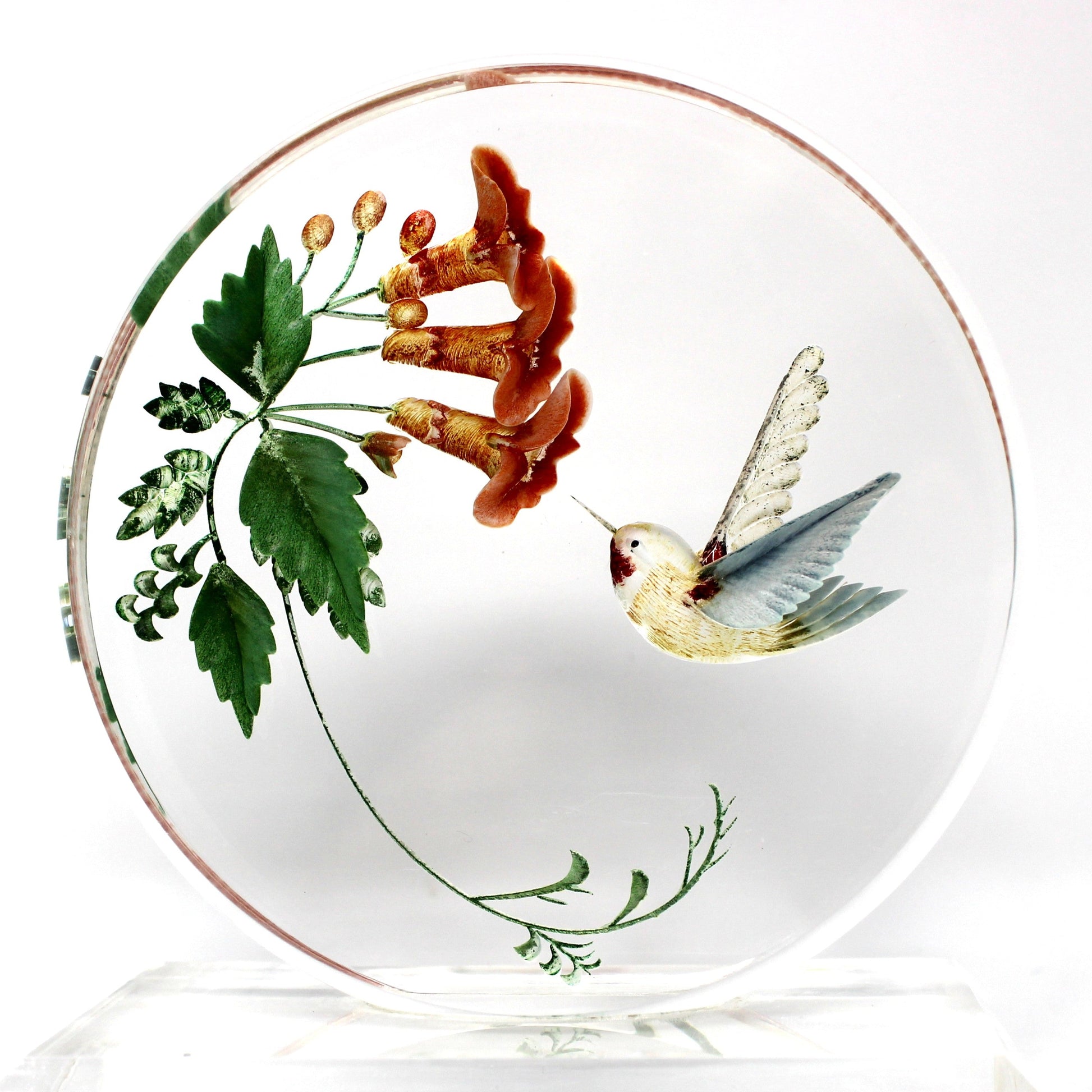 A round clear glass decorative plate featuring a painted illustration of a hummingbird and red-orange flowers with green leaves, displayed on a clear acrylic stand against a white background.