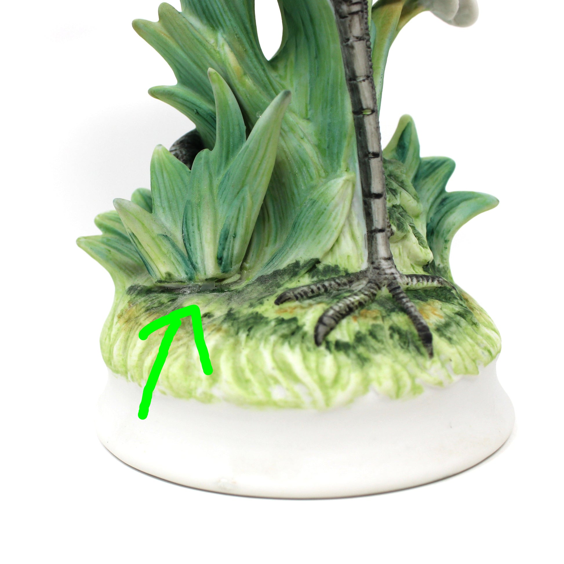A close-up of a ceramic bird figurine on a white base, showing green foliage, textured feathers, and a dark gray beak and legs against a plain white background.