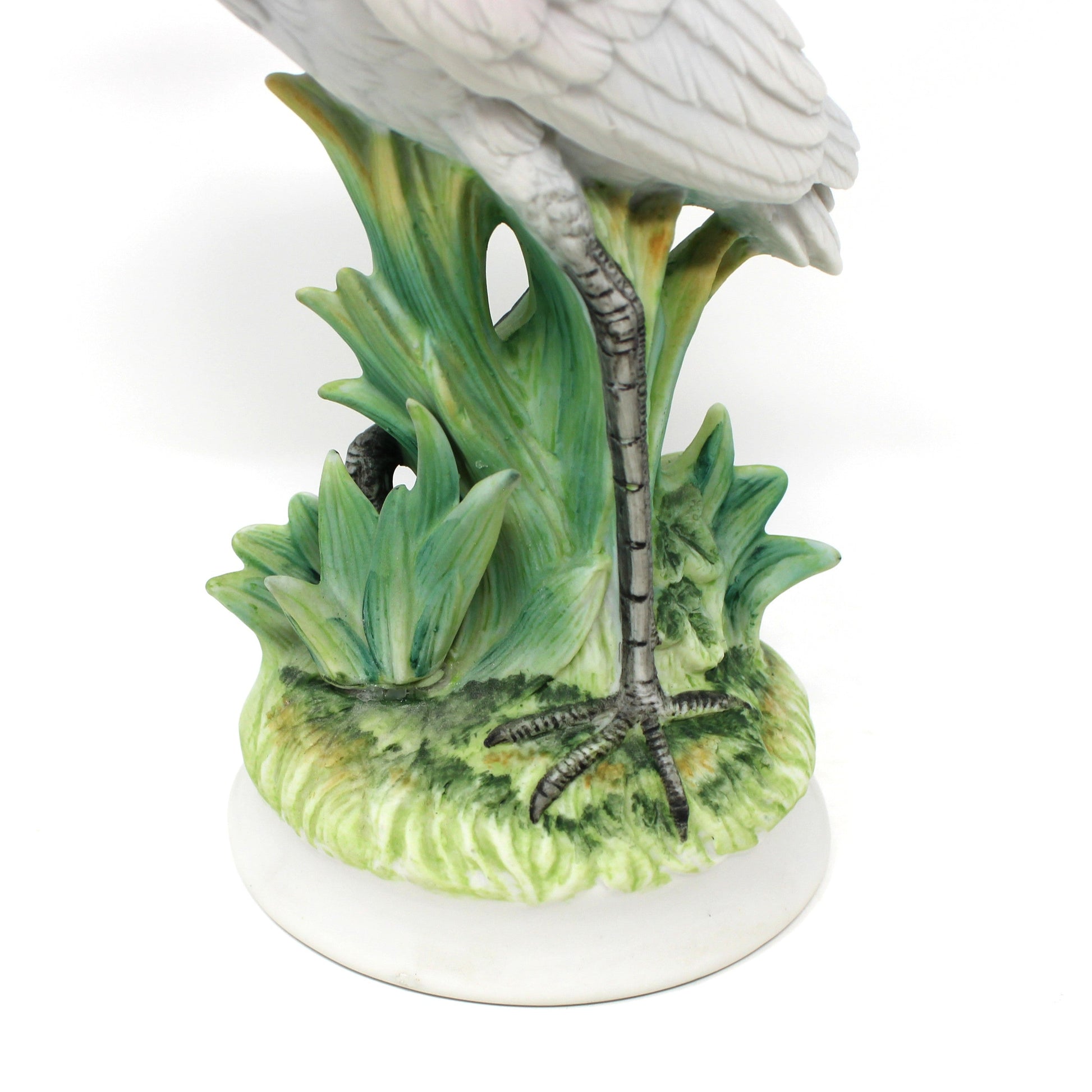 A ceramic heron figurine standing on green reeds over a round white base, photographed against a plain white background.
