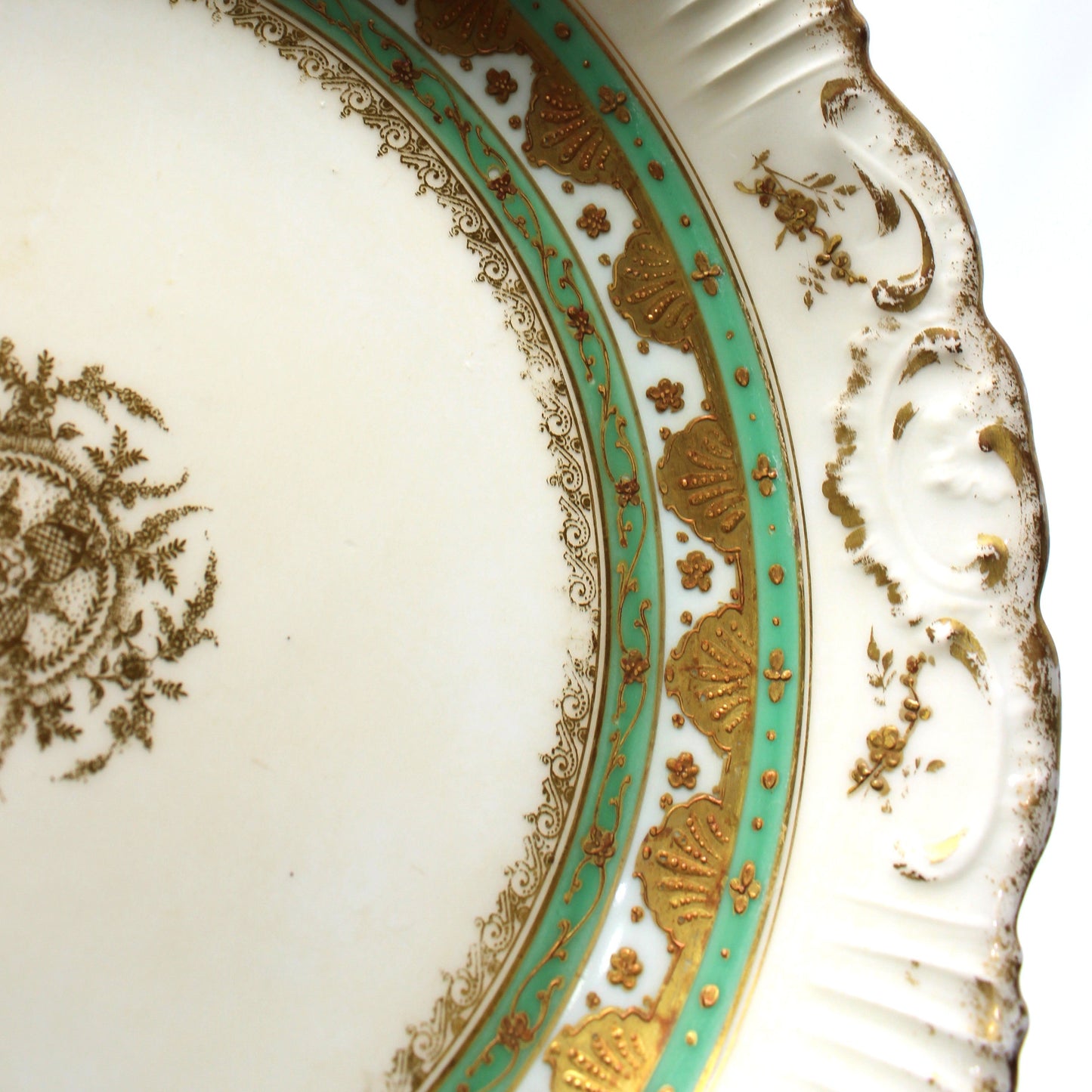 Close-up of an ornate white porcelain plate with a scalloped rim, featuring gold floral detailing, a turquoise and gold decorative band, and a gold medallion on the inner surface.