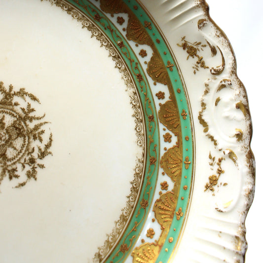 Close-up of an ornate white porcelain plate with a scalloped rim, featuring gold floral detailing, a turquoise and gold decorative band, and a gold medallion on the inner surface.