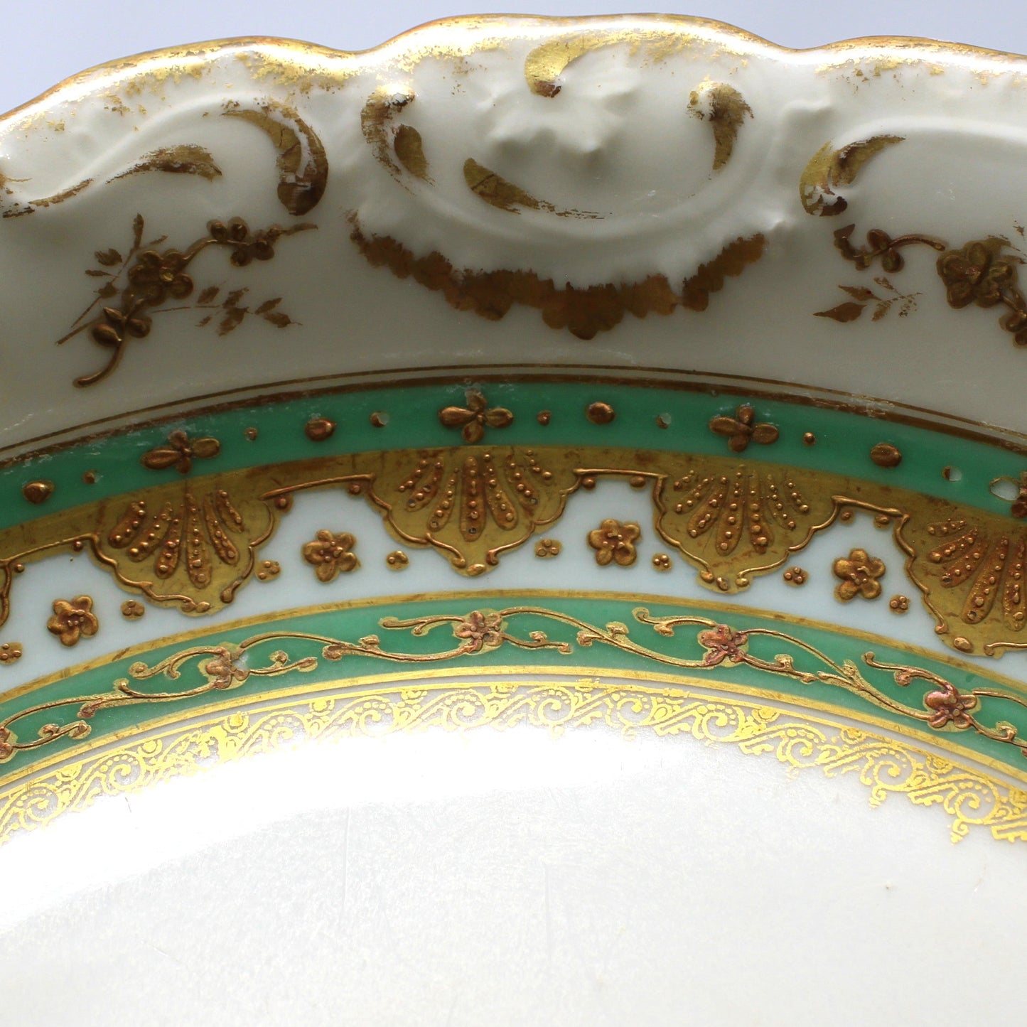 Close-up of an ornate white porcelain plate with a scalloped rim, decorated with gold scrollwork and a green band featuring gold fan motifs.