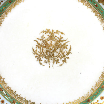 An ornate white porcelain bowl with a scalloped rim, decorated with intricate gold filigree, a central gold crest, and concentric rings of green and gold detailing.