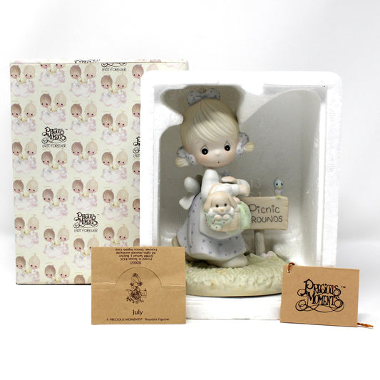 A vintage Precious Moments figurine titled July, displayed next to its original patterned gift box and two brown card inserts plus the foam packaging.